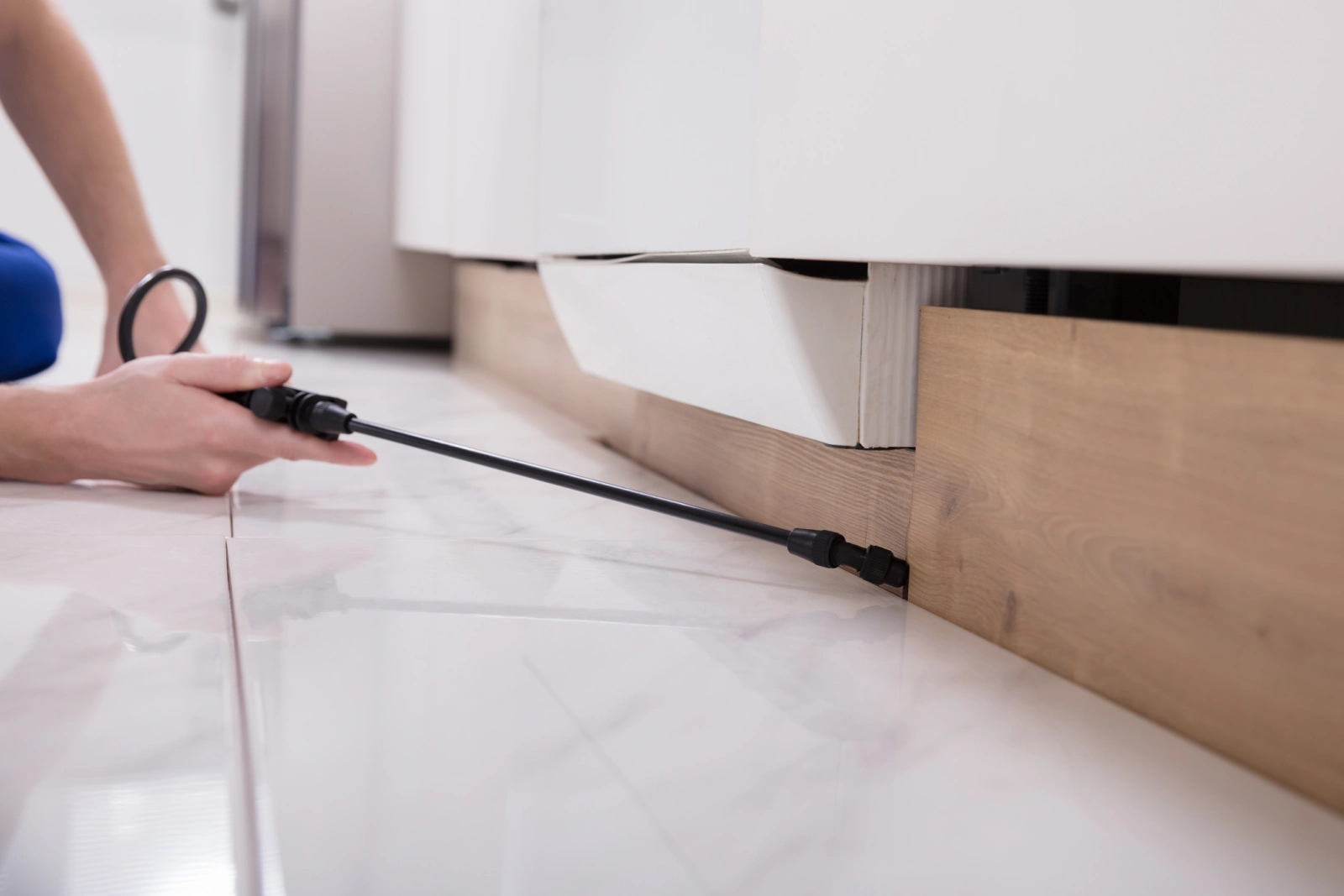 A close-up of a pest control technician's hand, spraying cockroach treatment on a baseboard.